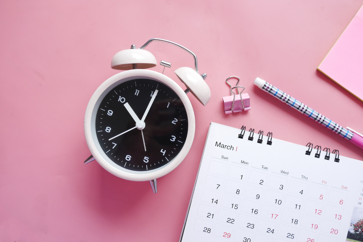 A pink desk on which is a classic alarm clock, pen, pink clip and a calendar.