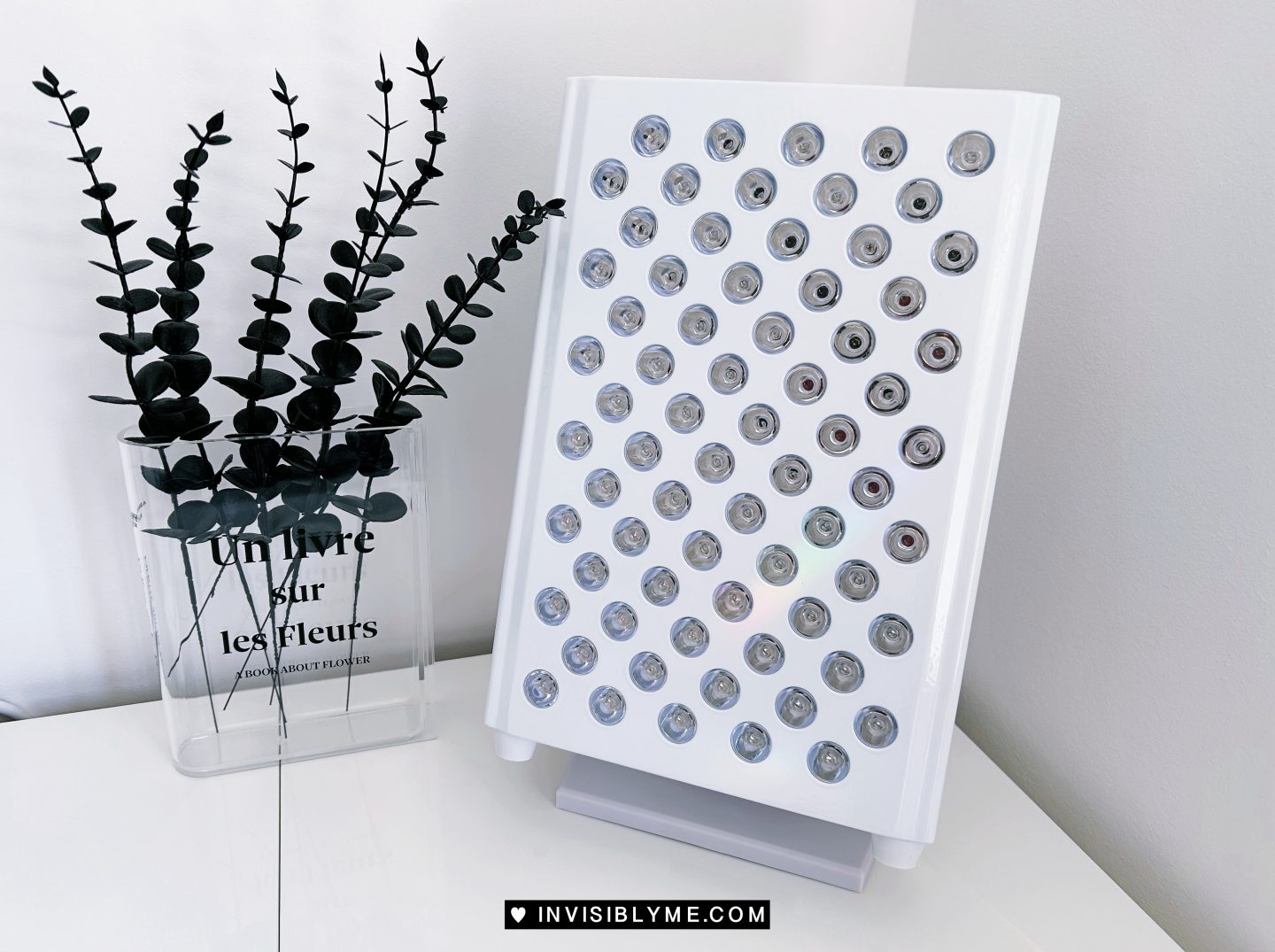 The Vital Red Light device on a white worktop against a white background. To the left is a clear perspex vase shaped like a book, with black leafy-flowers inside.